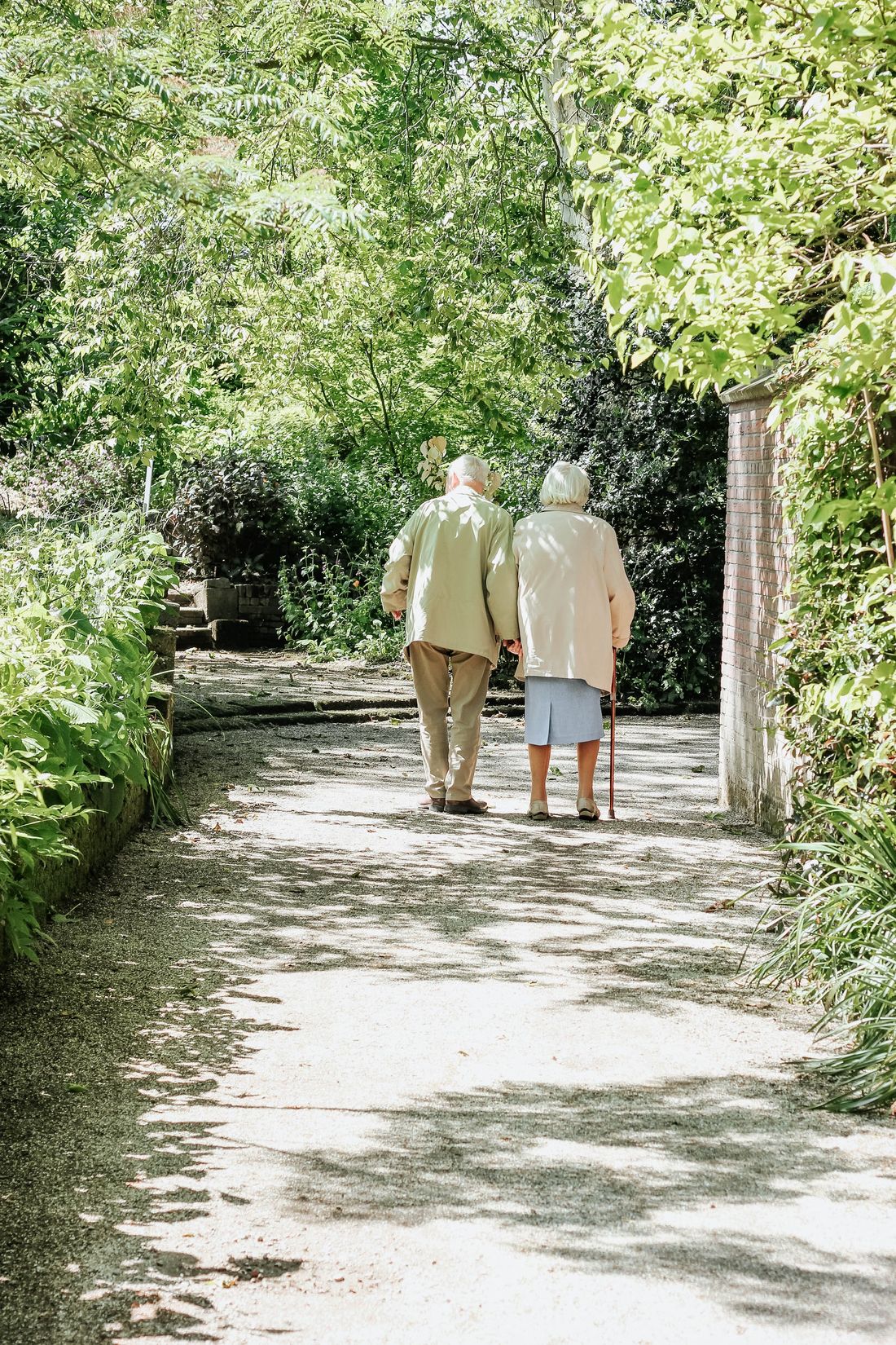An elderly couple walking arm in arm along a leafy garden path
